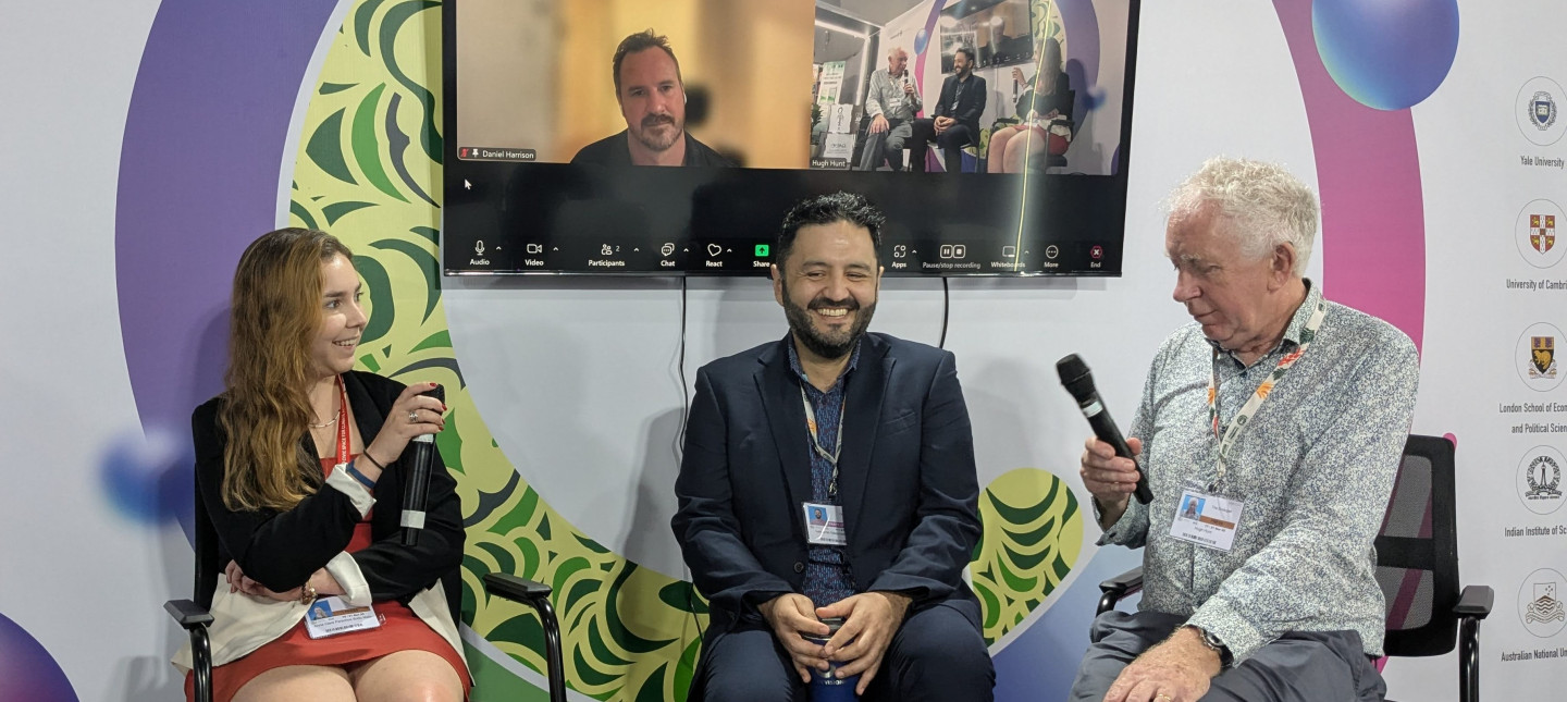 Three people with microphones, with one person behind on Zoom