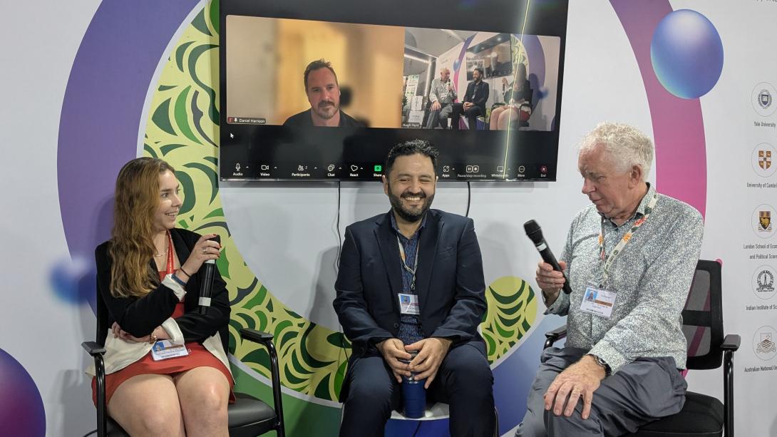 Three people with microphones, with one person behind on Zoom